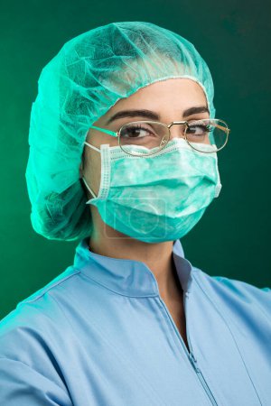 Photo for Doctor in blue lab coat, green operating theater cap and facial surgical mask looks serious through his eyeglasses, isolated on green background - Royalty Free Image