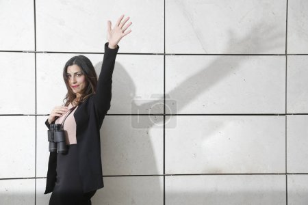 Foto de La mánager morena, vestida con traje de negocios negro, sostiene los prismáticos en la mano para mirar hacia otro lado, aislada sobre fondo de pared de azulejos rectangulares blancos - Imagen libre de derechos