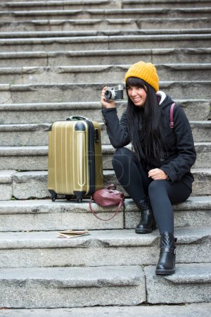 Téléchargez les photos : Jeune touriste en vêtements d'hiver assis sur les marches avec le chariot à proximité, prend une photo - en image libre de droit
