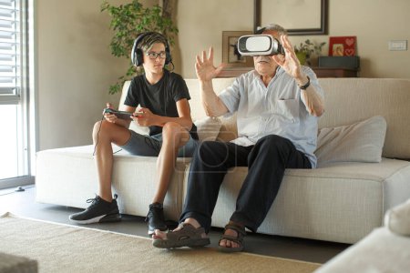 Téléchargez les photos : Grand-père et petit-fils jouent à des jeux électroniques assis sur le canapé dans le salon - en image libre de droit