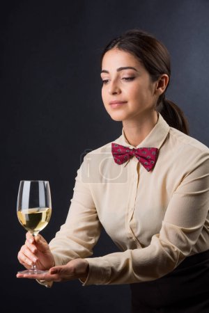 Foto de Hermosa y elegante camarera con camisa y pajarita roja está listo para ofrecer una copa de vino a un cliente, aislado sobre un fondo negro - Imagen libre de derechos