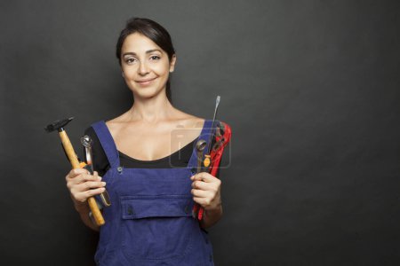 Photo for Brunette woman with mechanic work tools in hand and blue overalls makes face amused, isoalta on black background - Royalty Free Image