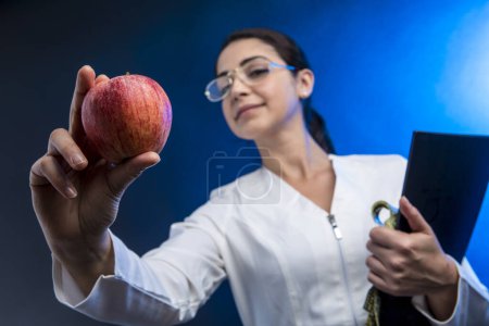Téléchargez les photos : Le diététicien en manteau blanc tient dans sa main un dossier noir et une pomme montrant fièrement, isolée sur un fond bleu - en image libre de droit