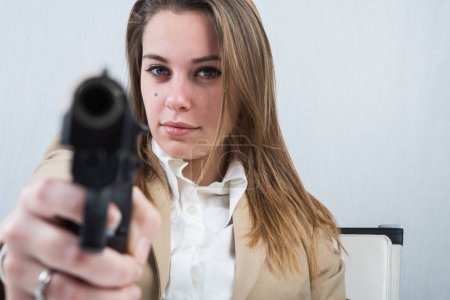 Foto de Chica rubia vestida con una chaqueta beige está sentada en su puesto de trabajo ella está sosteniendo un arma en su mano lista para usarlo - Imagen libre de derechos