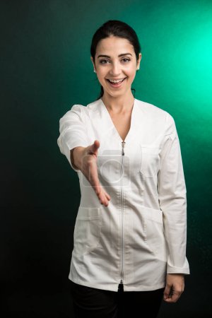 Photo for Female doctor in a white coat holds out her hand as a sign of knowledge and welcome to another hypothetical person, isolated on a green background - Royalty Free Image