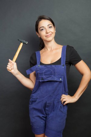 Photo for Beautiful girl with worker work tools in hand and blue overalls gurada curiously, isolated on black background - Royalty Free Image
