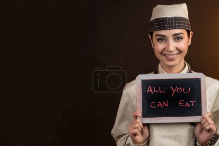 Photo for Beautiful and elegant chef in uniform holds a blackboard in his hand with "All you can eat" written on it, isolated on orange background - Royalty Free Image