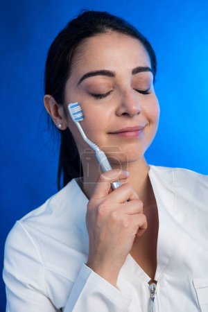 Photo for Girl in a white coat carries a toothbrush near her face, isolated on a blue background - Royalty Free Image
