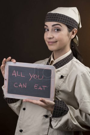 Photo for Beautiful and elegant chef in uniform holds a blackboard in his hand with "All you can eat" written on it, isolated on orange background - Royalty Free Image