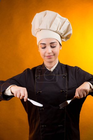 Téléchargez les photos : Cuisinier souriant avec chapeau de chef tient une fourchette et un couteau à la main, isolé sur fond orange - en image libre de droit