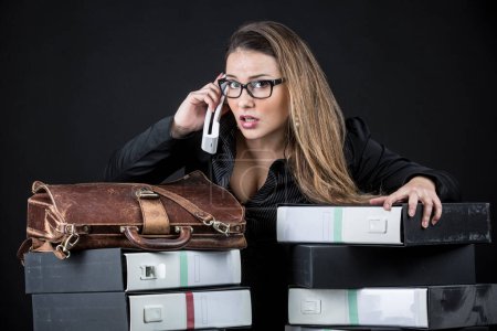 Photo for Blonde secretary with eyeglasses, dressed in black and desperate and worried amidst all the messy documents in her office, isolated on black background - Royalty Free Image