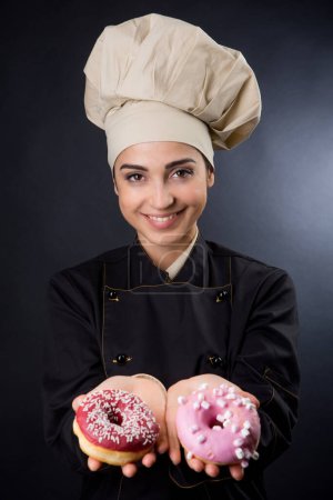 Foto de Cocinero sonriente muestra muy bien rosquillas de colores, aislado sobre fondo negro - Imagen libre de derechos