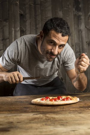 Téléchargez les photos : Un homme aux cheveux bruns barbu est sur le point de manger une pizza aux tomates cerises, isolé sur fond de bois - en image libre de droit