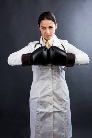 Téléchargez les photos : Femme médecin en manteau blanc et gants de boxe A l'expression de quelqu'un qui ne veut pas abandonner, isolé sur fond gris - en image libre de droit