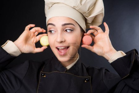 Foto de Bonito cocinero con sombrero beige y abrigo negro utiliza 2 macarrones como pendientes - fondo negro - Imagen libre de derechos