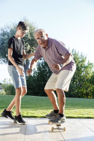 Photo for Teenage grandson teaches his Elder grandfather to skateboard in the garden - Royalty Free Image