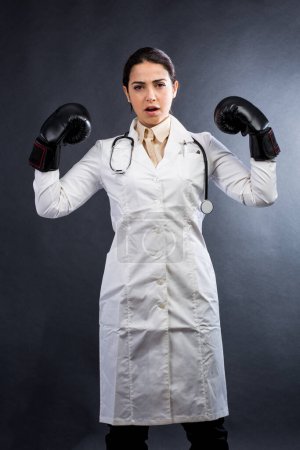 Foto de Doctora en bata blanca y guantes de boxeo Tiene la expresión de alguien que no quiere rendirse, aislado sobre fondo gris - Imagen libre de derechos