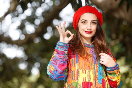 Photo for Portrait of beautiful white girl with colorful sweater and red headphone, isolated on nature background making the ok sign with her fingers - Royalty Free Image