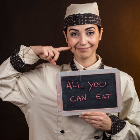 Foto de Hermoso y elegante chef en uniforme sostiene una pizarra en la mano con "Todo lo que puedes comer" escrito en él, aislado sobre fondo naranja - Imagen libre de derechos
