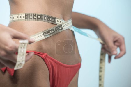 Foto de Chica en bikini rojo gira una cinta métrica alrededor de su cintura para tomar medidas exactas, aislado sobre fondo claro - Imagen libre de derechos