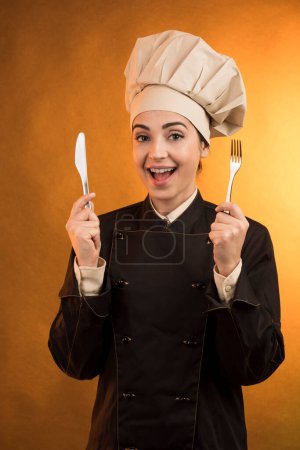 Foto de Cocinero sonriente con sombrero de chef sostiene un tenedor y un cuchillo en la mano, aislado sobre fondo naranja - Imagen libre de derechos