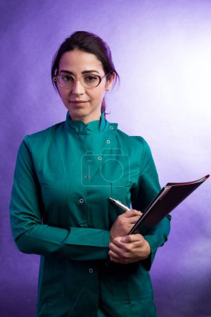 Foto de Doctora en bata de laboratorio verde sosteniendo una carpeta negra y gafas mientras mira severamente, aislada sobre fondo púrpura - Imagen libre de derechos