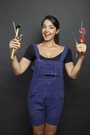Téléchargez les photos : Femme brune avec des outils de travail mécanique en salopette bleue et à la main rend le visage amusé, isoalta sur fond noir - en image libre de droit