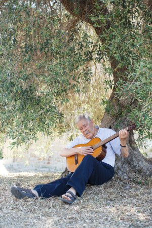 Téléchargez les photos : Homme de quatre-vingts ans mélancolique jouant une mélodie assise à l'ombre d'un vieil olivier à la campagne - en image libre de droit
