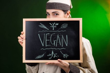 Photo for Uniformed cook holds a blackboard with "Vegan" written on it, behind which she is hiding, isolated on a green background. - Royalty Free Image