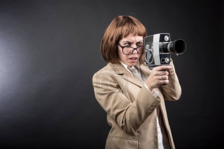 Foto de Chica con gafas, pelo bob y chaqueta beige, utiliza una vieja cámara de cine mirándola con expresión divertida de incertidumbre, aislado sobre fondo negro - Imagen libre de derechos