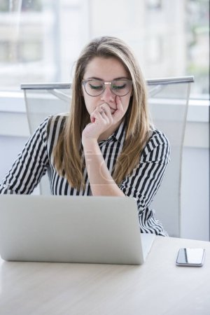 Téléchargez les photos : Gérant blonde avec des lunettes et chemise rayée travaille dans le bureau assis devant son lieu de travail - en image libre de droit