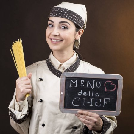 Foto de Cocinera sosteniendo pizarra con "Chef 's Menu" escrito en su mano sosteniendo algunos espaguetis crudos, aislados sobre fondo marrón - Imagen libre de derechos