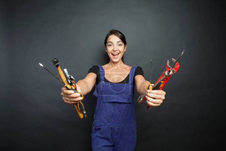 Téléchargez les photos : Femme brune avec des outils de travail mécanique en salopette bleue et à la main rend le visage amusé, isoalta sur fond noir - en image libre de droit