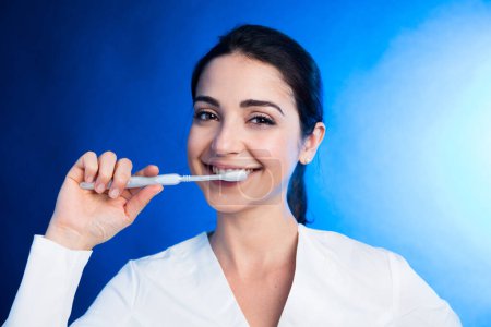 Foto de Chica en un abrigo blanco muestra cómo cepillarse bien los dientes con un cepillo de dientes, aislado sobre un fondo azul - Imagen libre de derechos