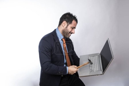 Photo for Dark-haired man with beard, dressed in shirt suit, jacket and orange tie, takes a laptop in his hand with hammer, isolated on white background - Royalty Free Image