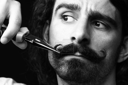 Téléchargez les photos : Homme aux cheveux bruns utilise des pinces pour fixer sa moustache - isolé sur fond noir - en image libre de droit