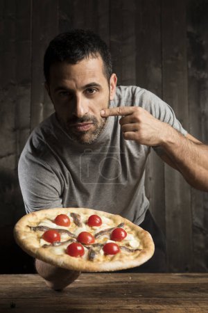 Foto de Hombre de pelo oscuro con barba está a punto de comer una pizza con tomates cherry, aislado sobre fondo de madera - Imagen libre de derechos