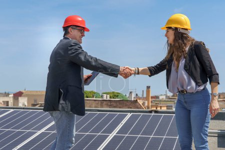 Foto de Pareja de ingenieros con cascos protectores estrechando las manos de acuerdo cerca de un sistema fotovoltaico en el techo de una casa. - Imagen libre de derechos