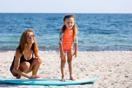 Téléchargez les photos : Mère en maillot de bain enseigne les bases du surf à sa fille sur une plage balnéaire. - en image libre de droit