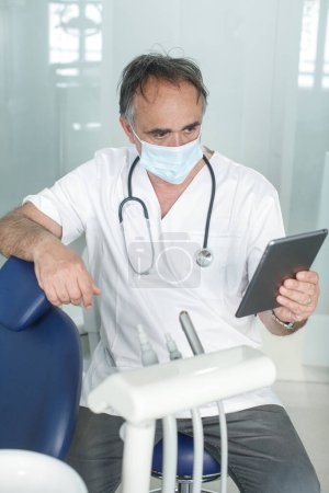 Téléchargez les photos : Médecin de cinquante ans avec masque chirurgical en manteau blanc, regarde un comprimé - en image libre de droit