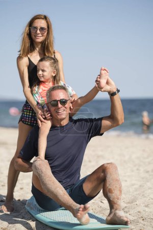 Téléchargez les photos : Famille joyeuse composée de père, mère et fille s'amuse à jouer avec une planche de surf sur la plage à la mer - en image libre de droit