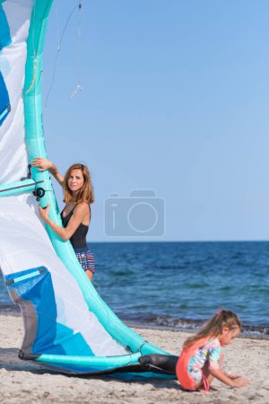Foto de Mujer blanca con vela kitesurf mira a la distancia. La hijita juega con arena en primer plano. - Imagen libre de derechos