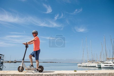 Älterer Mann in orangefarbenem Trikot und Schutzhelm fährt auf seinem Elektroroller vor dem Yachthafen, isoliert auf Himmelshintergrund 