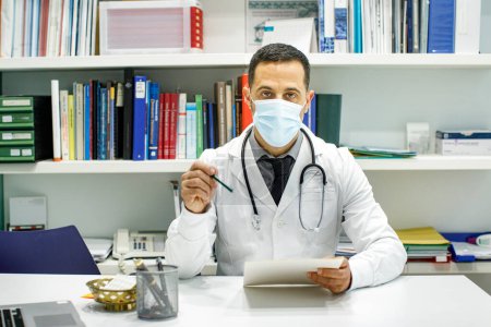 Foto de Médico de pelo negro con una bata blanca lee datos sentados en el escritorio de su consultorio médico - Imagen libre de derechos