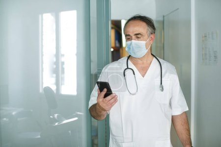 Téléchargez les photos : Médecin de 50 ans en manteau blanc et masque chirurgical regarde son téléphone portable dans une salle d'hôpital - en image libre de droit