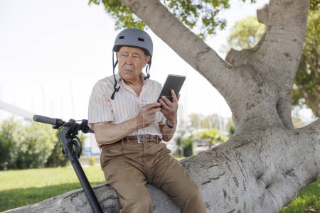 Foto de Anciano con un casco protector utiliza su tableta para navegar por Internet sentado junto a su scooter eléctrico en un parque - Imagen libre de derechos