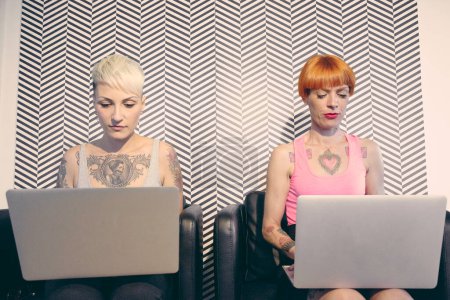 Foto de Dos chicas jóvenes tatuadas trabajan en las computadoras portátiles - Imagen libre de derechos