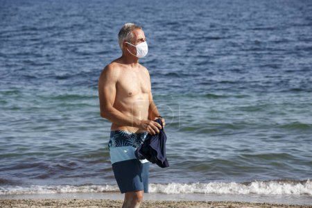 Photo for Shirtless fifty-year-old man stands on the beach in a swimsuit while taking off his protective mask - Royalty Free Image