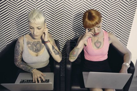 Téléchargez les photos : Deux jeunes filles tatouées travaillent sur les ordinateurs portables - en image libre de droit
