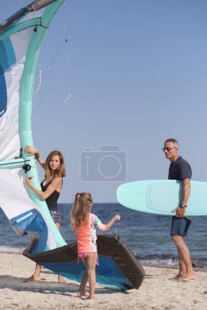 Photo for Family consisting of father, mother and a daughter prepares kitesurfing on the beach ready to use it at sea. - Royalty Free Image